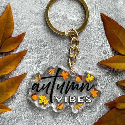 Keychain with 'autumn vibes' text surrounded by autumn leaves on a gray surface.