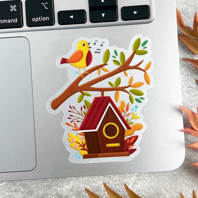 Sticker of a birdhouse and bird on a branch with leaves on a laptop.