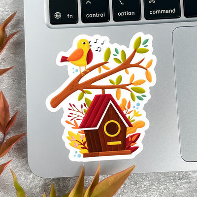 Sticker of a birdhouse with a bird on a branch on a laptop keyboard.