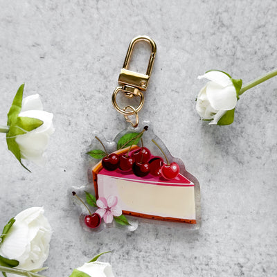 Keychain shaped like a slice of cake with cherries on a gray surface with white flowers.