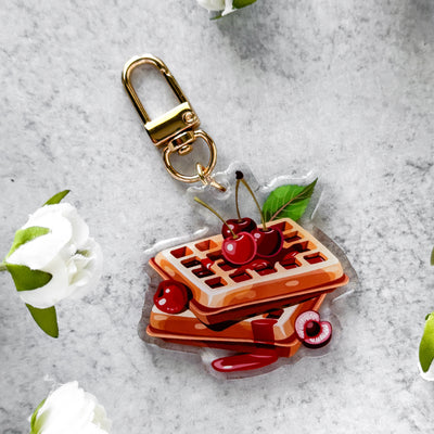 Waffle and cherry keychain on a gray surface with white flowers.