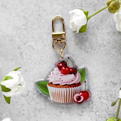 Cupcake keychain with pink frosting and cherries on a gray surface with white flowers.