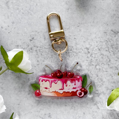 Keychain shaped like a cake with cherries on a gray surface with flowers.