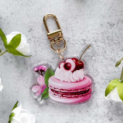 Keychain shaped like a pink macaron with a cherry on top, surrounded by flowers on a gray surface.