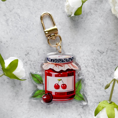 Keychain with a jar of red jam and cherries on a gray surface with white flowers.