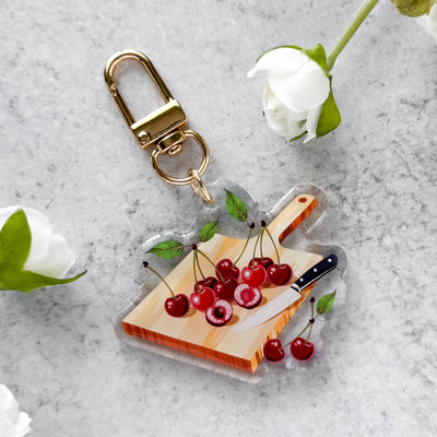 Keychain with a cutting board design featuring cherries and a knife on a gray surface with white flowers.
