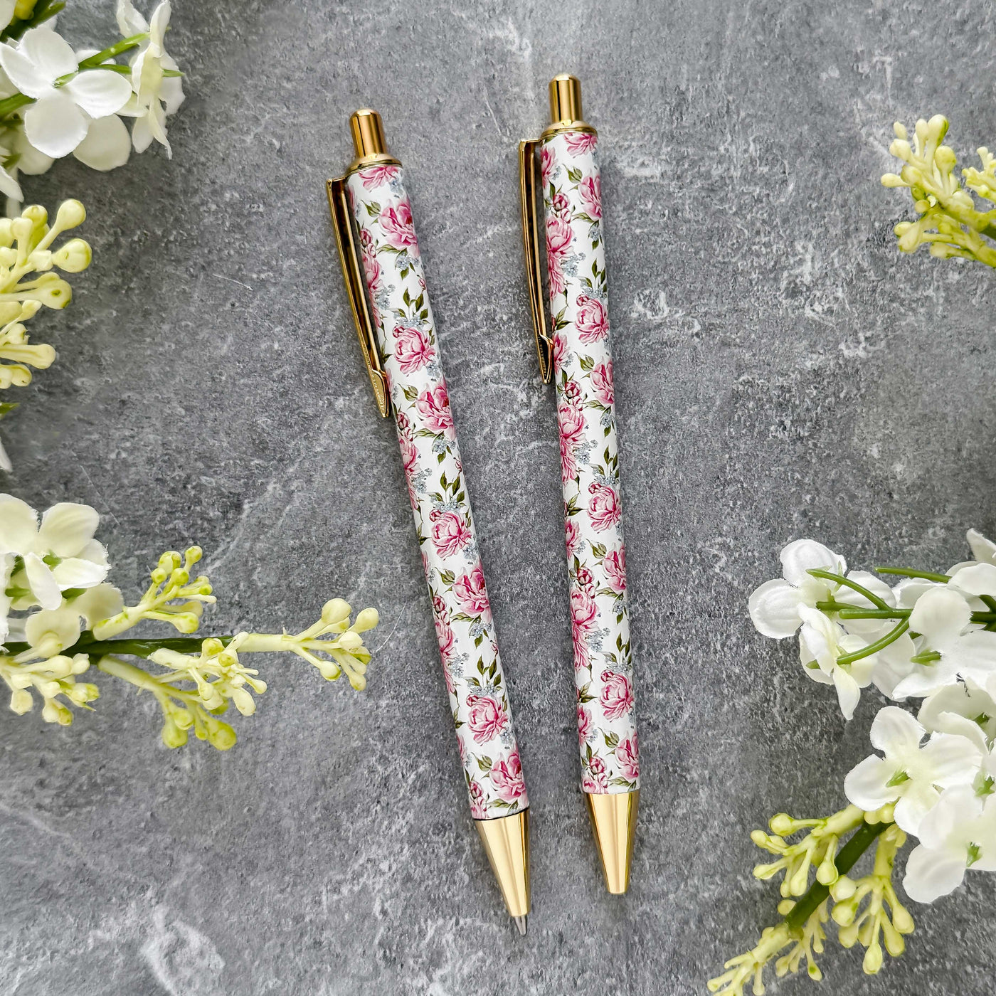 Two floral-patterned pens on a gray surface with white flowers around them.