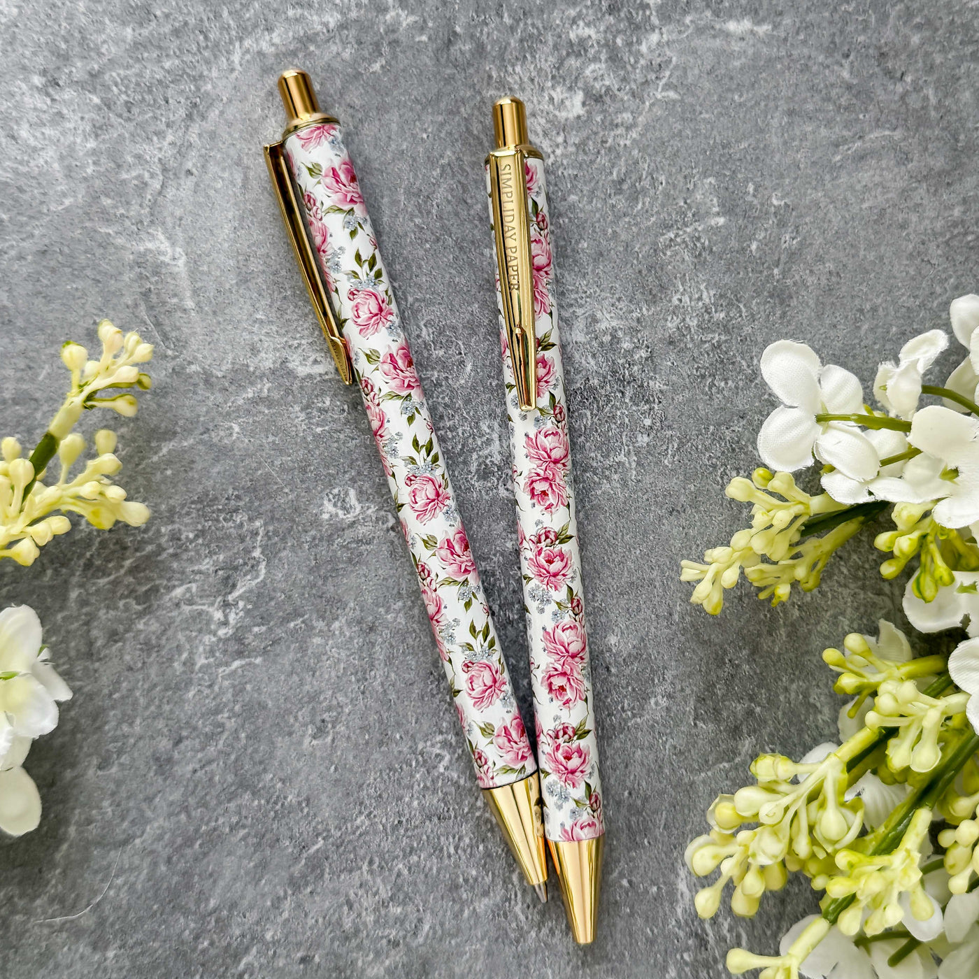 Two floral-patterned pens with gold accents on a gray surface with flowers around.