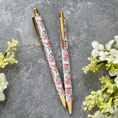Two floral-patterned pens with gold accents on a gray surface with flowers around.
