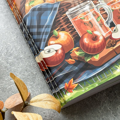 Notebook with autumn-themed cover featuring apples, leaves, and a basket on a gray surface.