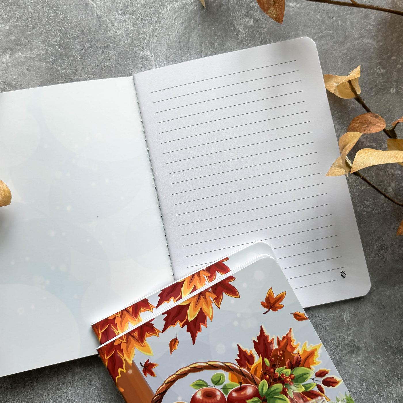 Open notebook with lined pages on a gray surface, accompanied by a decorative card with autumn leaves and fruits.