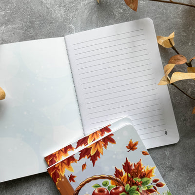 Open notebook with lined pages on a gray surface, accompanied by a decorative card with autumn leaves and fruits.