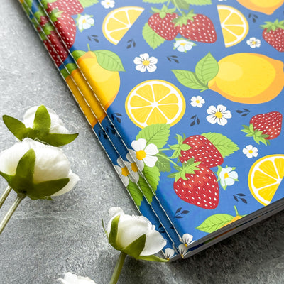 Notebook with a colorful fruit and floral pattern on a gray surface with white flowers.