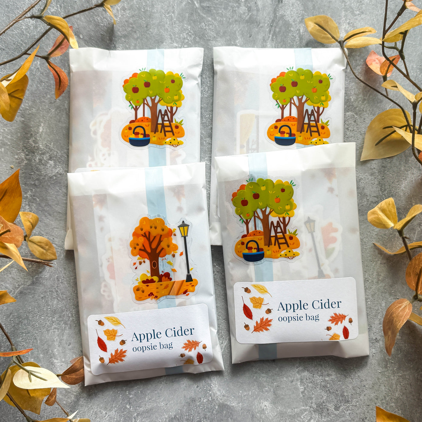 Four packages of 'Apple Cider' oopsie bags with autumn-themed designs on a gray surface.