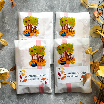 Four 'Autumn Cafe oopsie bag' packages with a tree design on a gray surface with autumn leaves.