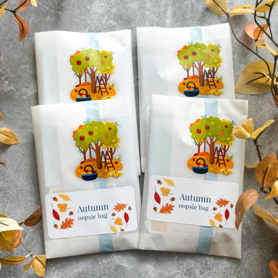 Four white bags with autumn-themed designs and text on a gray surface with decorative leaves.