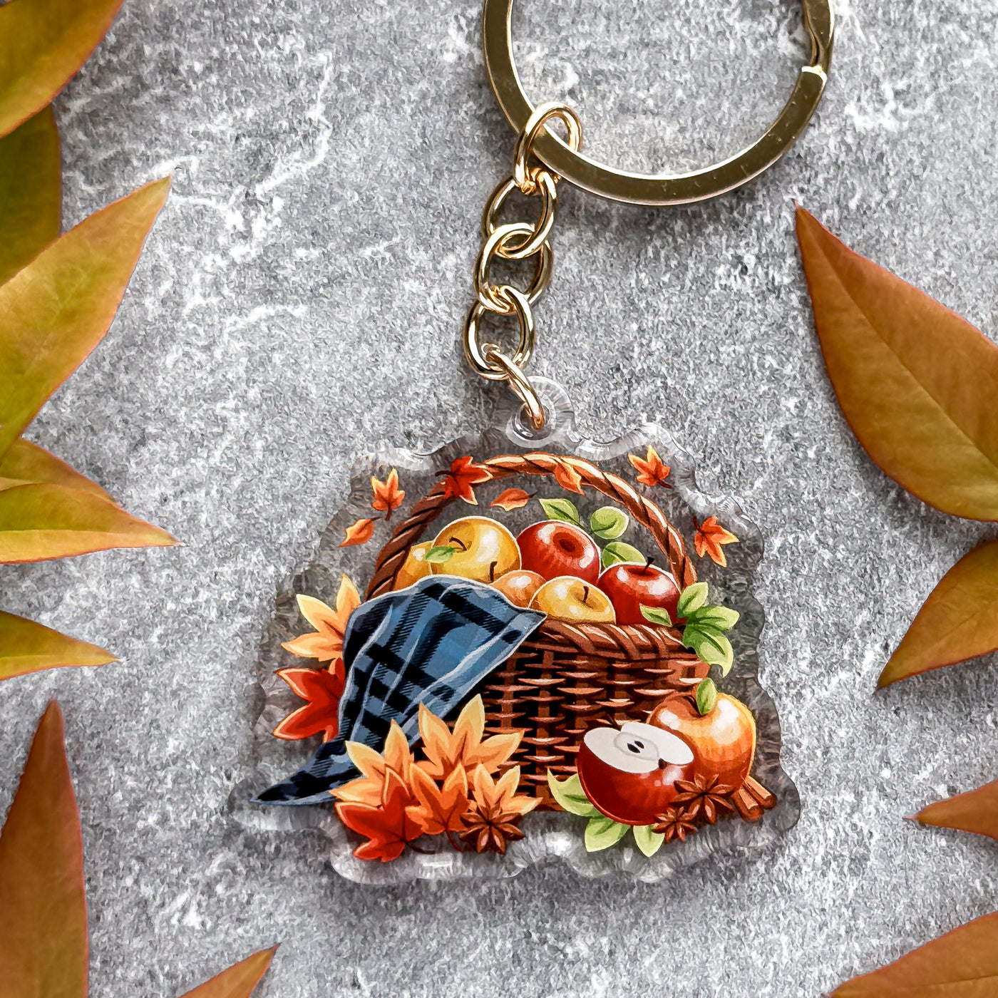 Keychain with a design of a basket of apples and leaves on a textured surface.