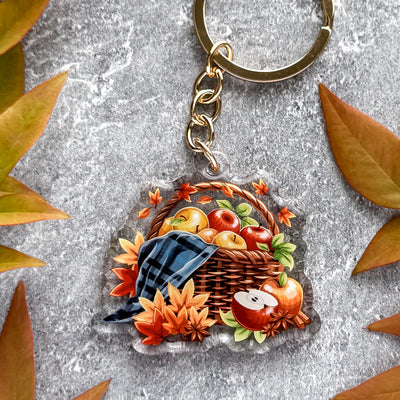 Keychain with a design of a basket of apples and leaves on a textured surface.